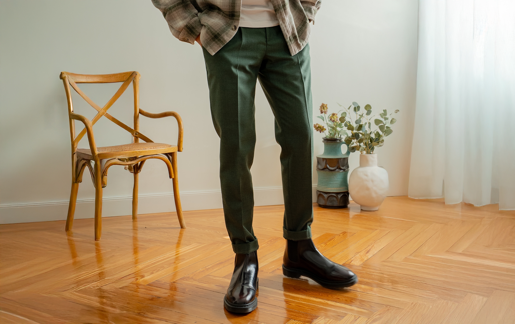 Un hombre de pie viste una playera beige y pantalones oscuros con un corte moderno. Lleva las manos en los bolsillos y usa tenis blancos. El fondo es de color gris claro, lo que resalta el atuendo.